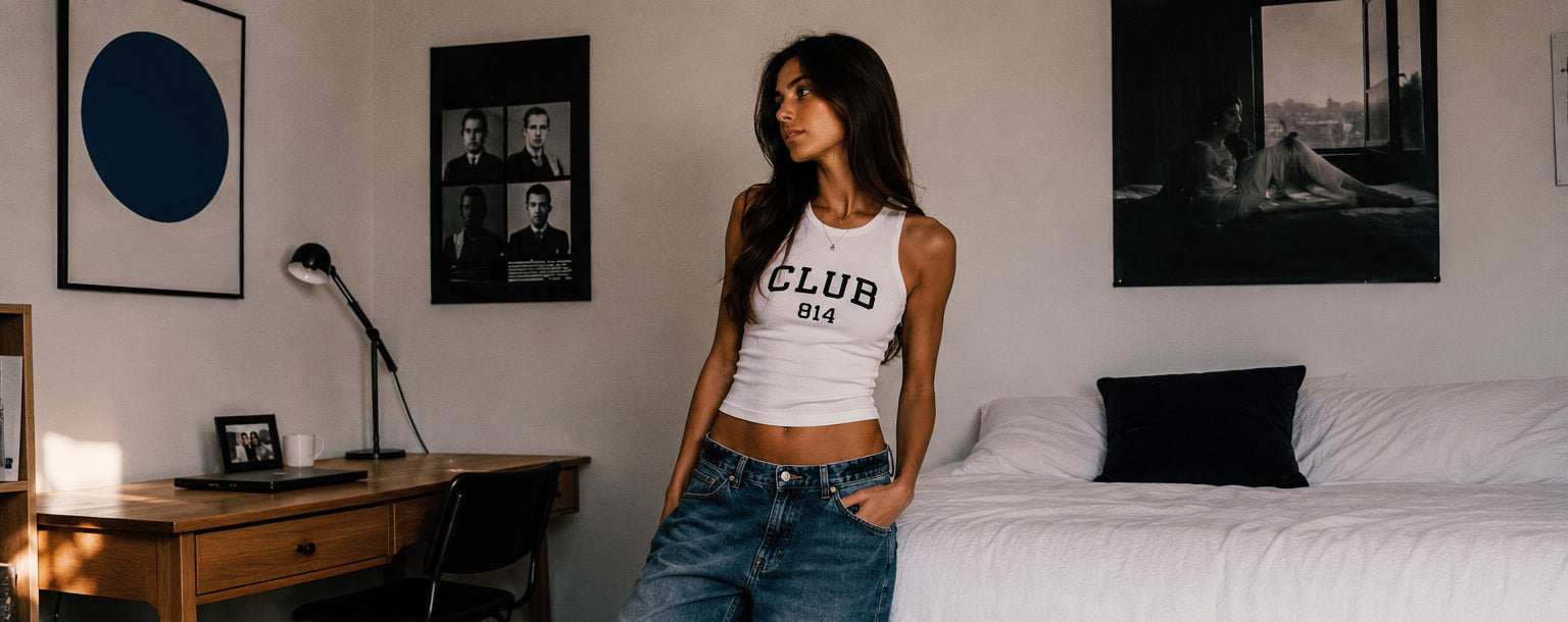Woman in a room with a desk and bed, wearing a 'CLUB 814' t-shirt and jeans.