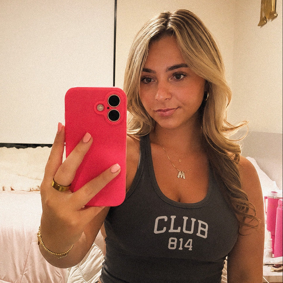A woman taking a mirror selfie in a bedroom, wearing a dark gray tank top with 'CLUB 814' printed in white. She is holding a bright pink smartphone.