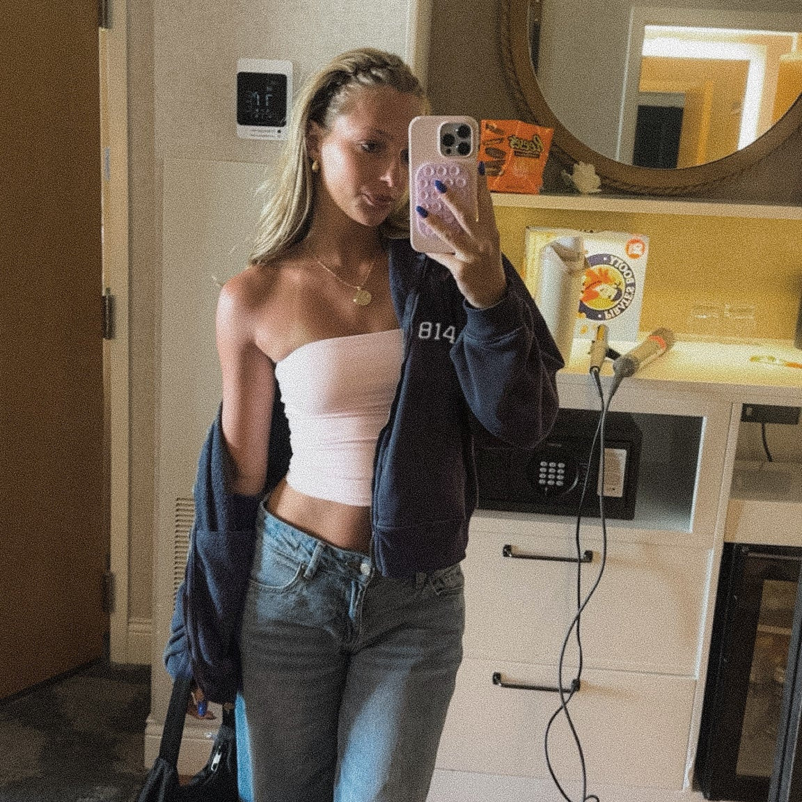 A college girl takes a mirror selfie in a dorm room, wearing a light pink strapless tube top, blue flare jeans, and a dark navy zip-up Club 814 hoodie draped off her shoulder.