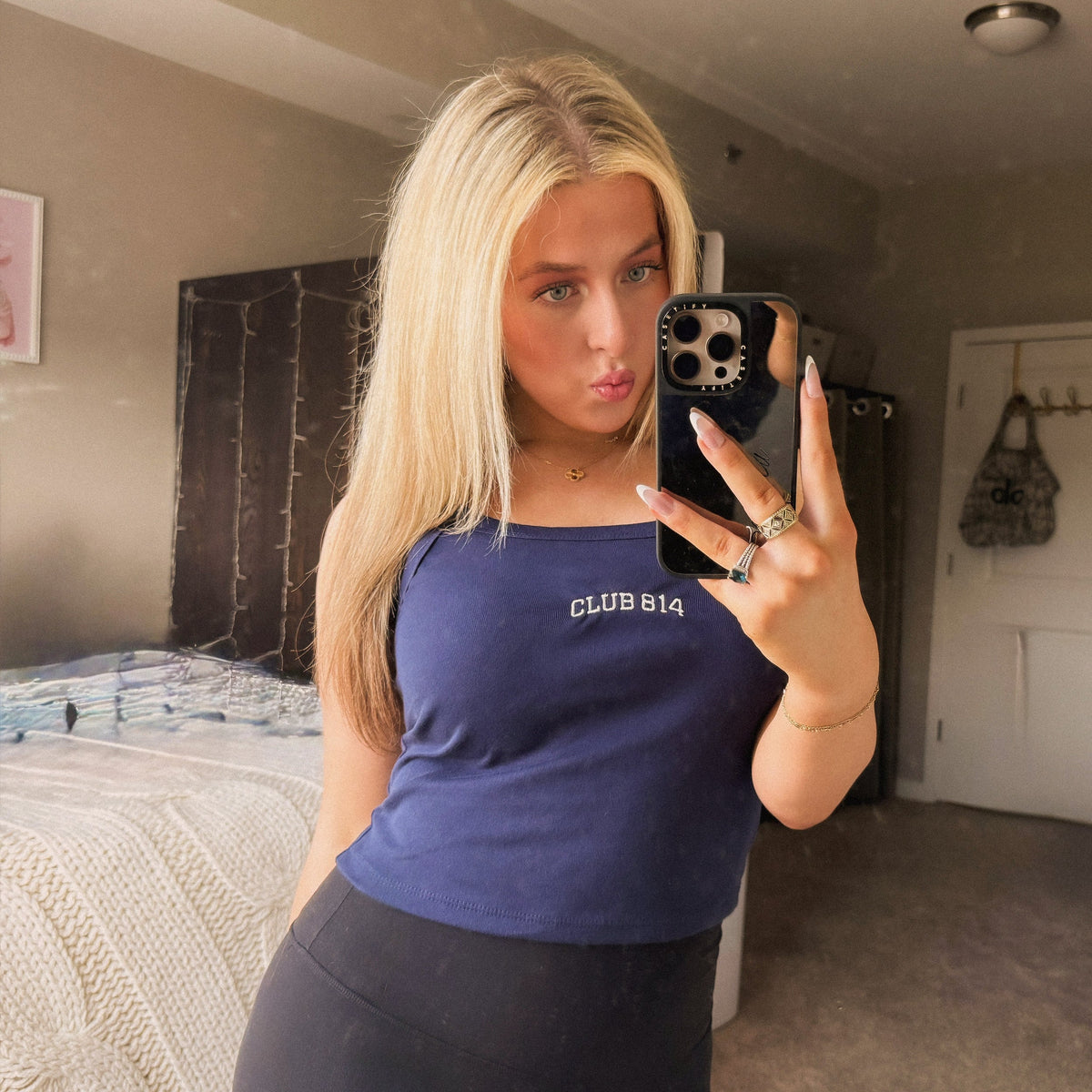 A woman taking a mirror selfie in a bedroom, wearing a navy blue cropped tank top with 'CLUB 814' printed on the chest and black leggings.