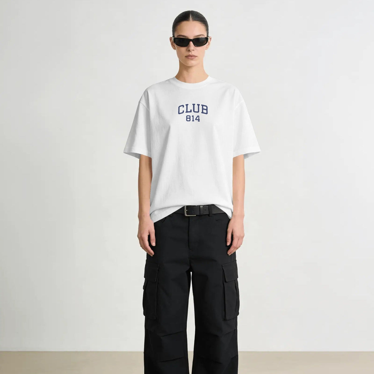 A model wearing a white oversized crewneck t-shirt with Club 814 on the front, paired with black cargo pants and black sunglasses, set against a plain light gray background.