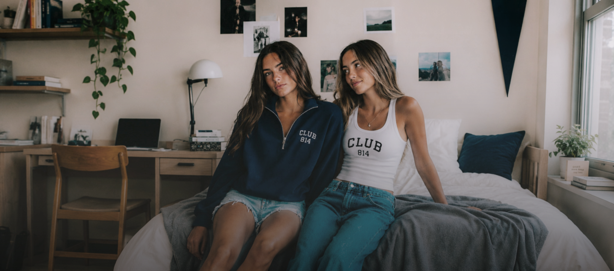 Two women sitting on a bed in a bedroom setting, wearing Club 814 brand apparel. One wears a navy blue zip-up hoodie and denim shorts, while the other wears a white tank top and teal wide-leg jeans.