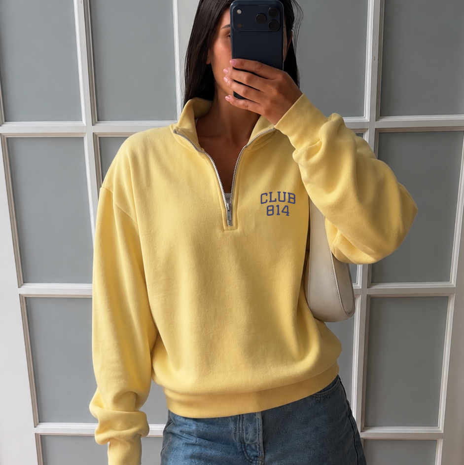 A woman takes a mirror selfie wearing a pale yellow quarter-zip sweatshirt with 'CLUB 814' printed on the chest, paired with denim shorts and yellow flip-flops, standing in front of a white paneled door.