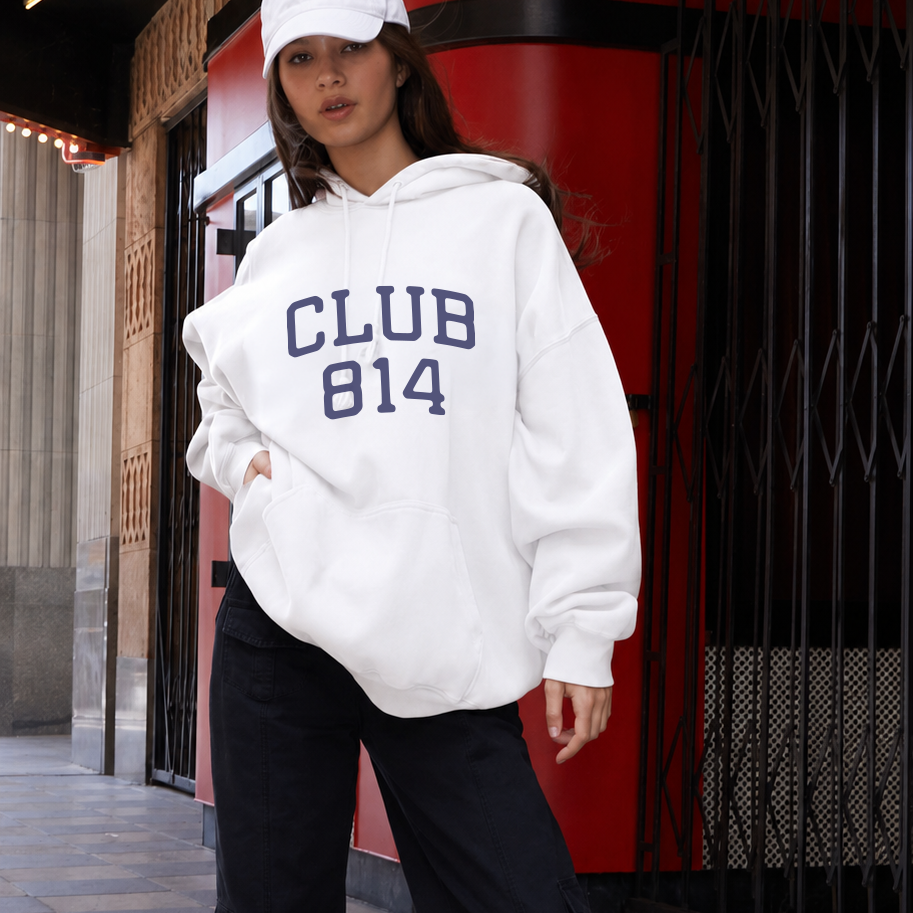 A woman wearing a white oversized hoodie with 'CLUB 814' printed in navy blue varsity lettering, paired with black wide-leg pants, a white cap, and white sneakers, standing on a checkered sidewalk in front of a red and black storefront.