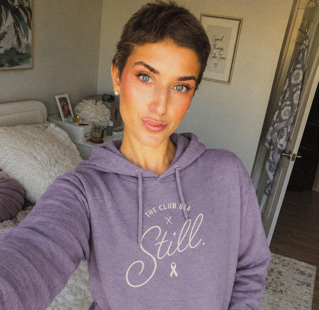 A woman takes a mirror selfie wearing a lavender purple hoodie with 'THE CLUB GIFT x Still.' printed in white script on the chest, paired with black leggings in a bedroom setting.