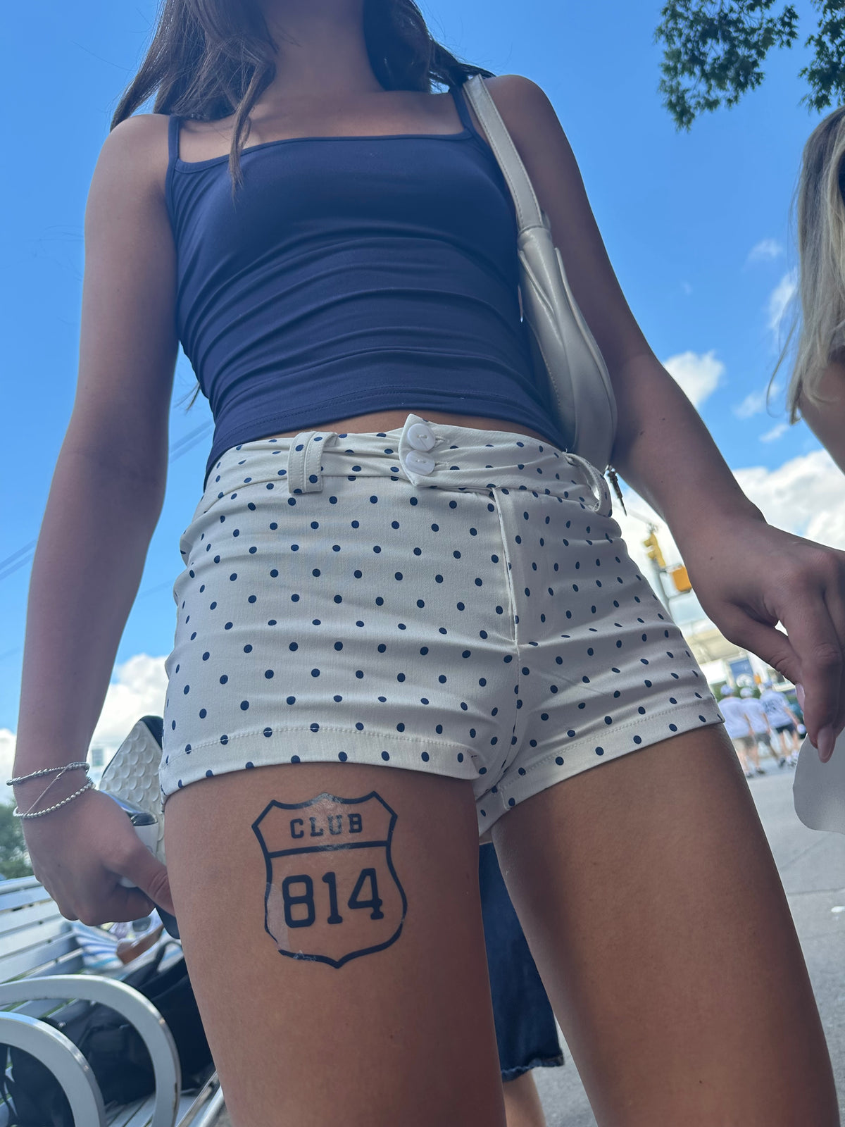 A person outdoors wearing white shorts with navy blue polka dots and a navy blue ribbed tank top. A black Route 66 style shield graphic is printed on the upper thigh reading 'CLUB 814'.