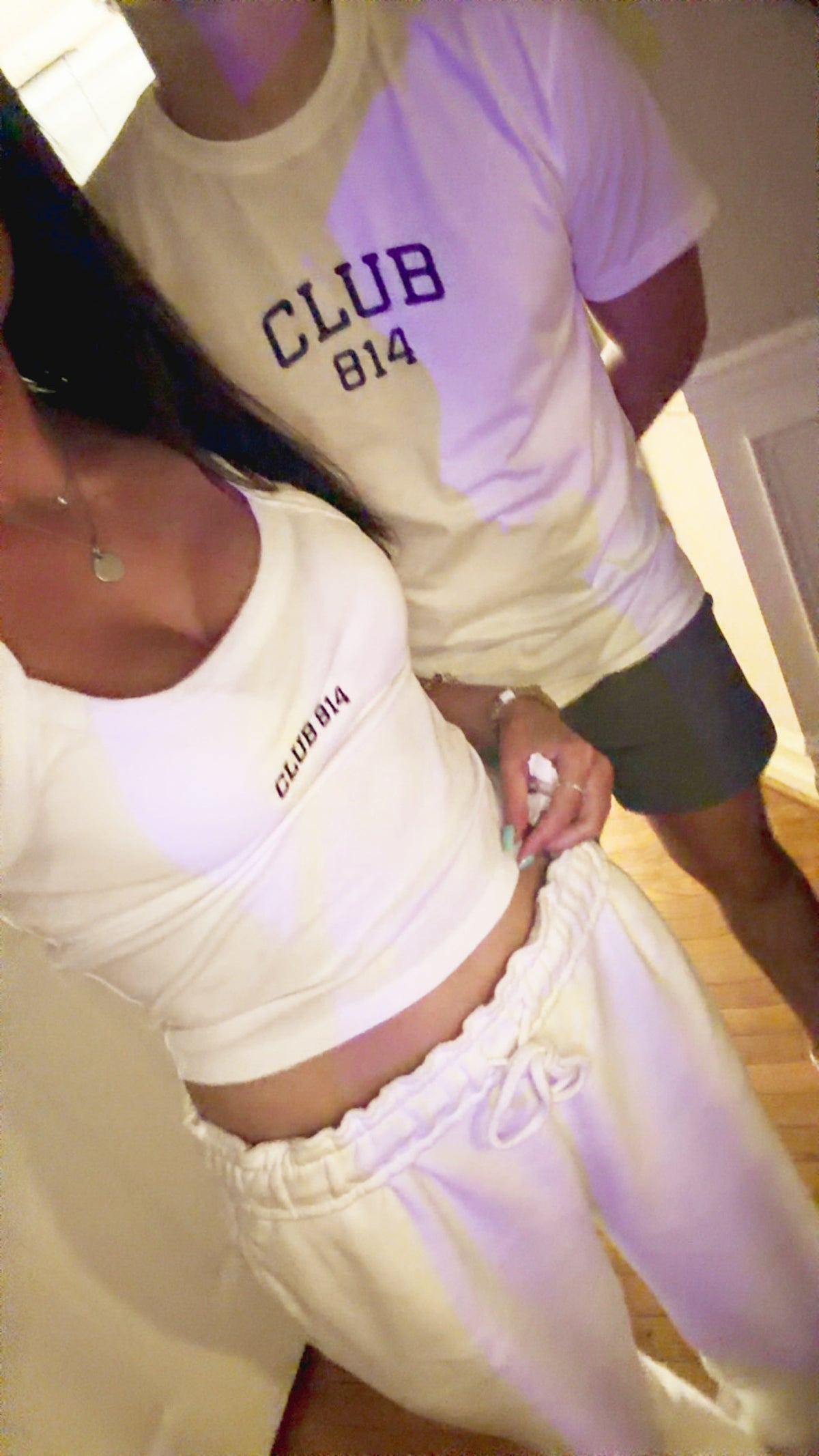 A couple wearing matching white t-shirts in a mirror selfie. The man's shirt is cream with purple side panels and reads 'CLUB 814' in purple.