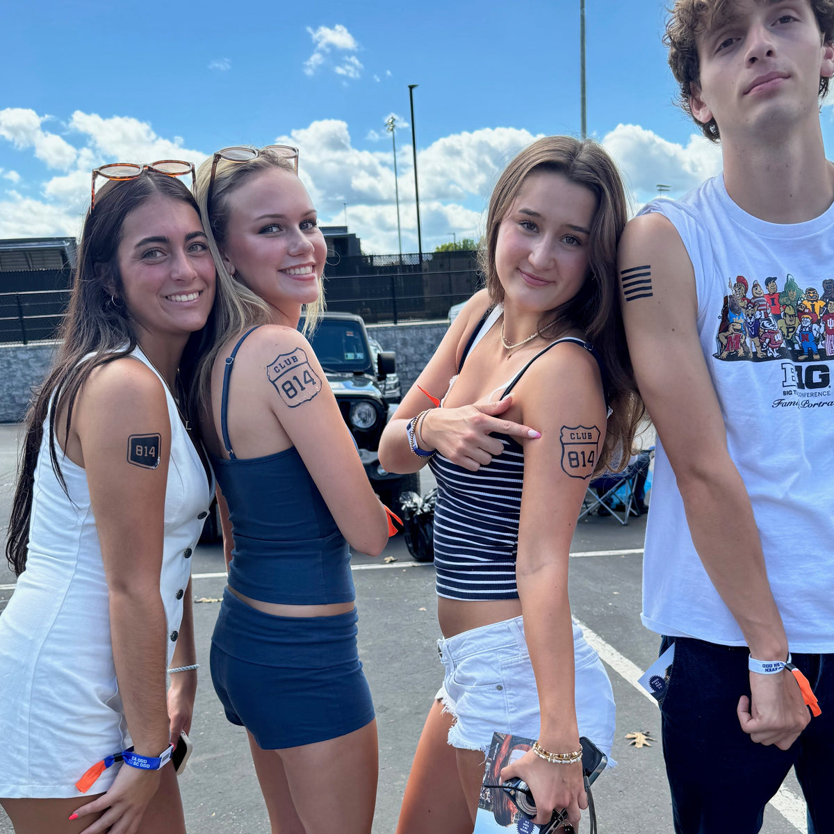 Four young adults posing together in an outdoor parking lot under a blue sky with clouds. They are wearing casual summer outfits, including tank tops, shorts, boots, and sneakers.