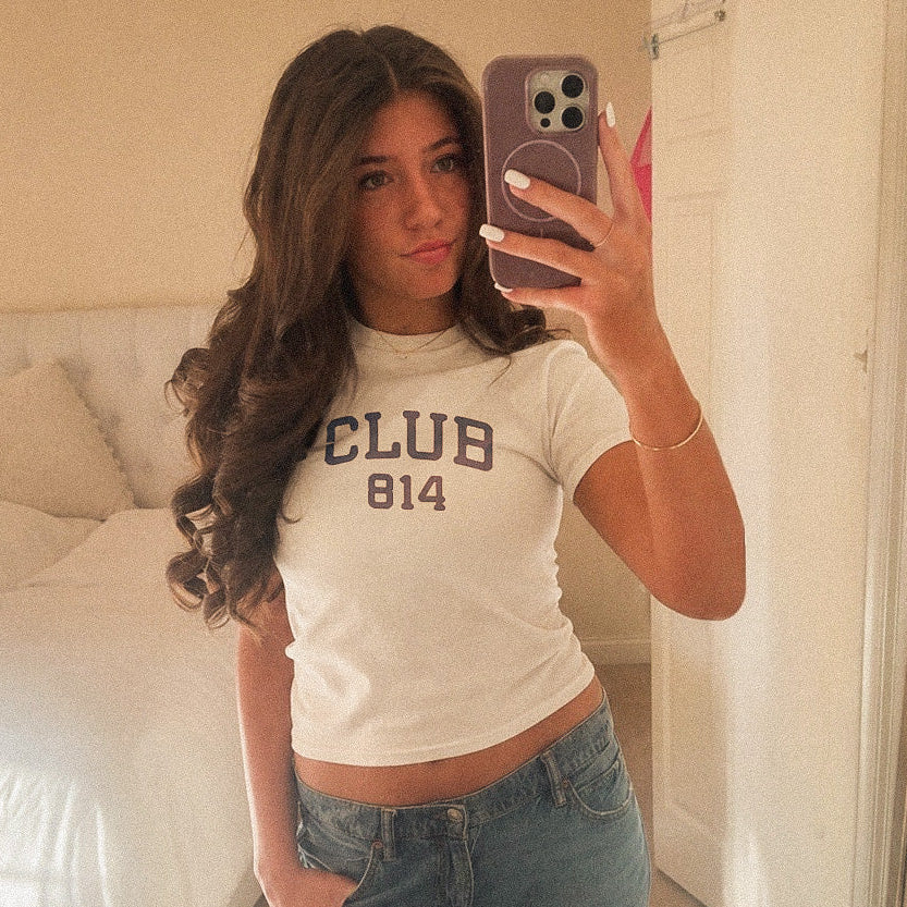 A woman takes a mirror selfie in a bedroom while wearing a fitted off-white crop top with 'CLUB 814' printed in dark lettering and blue jeans.