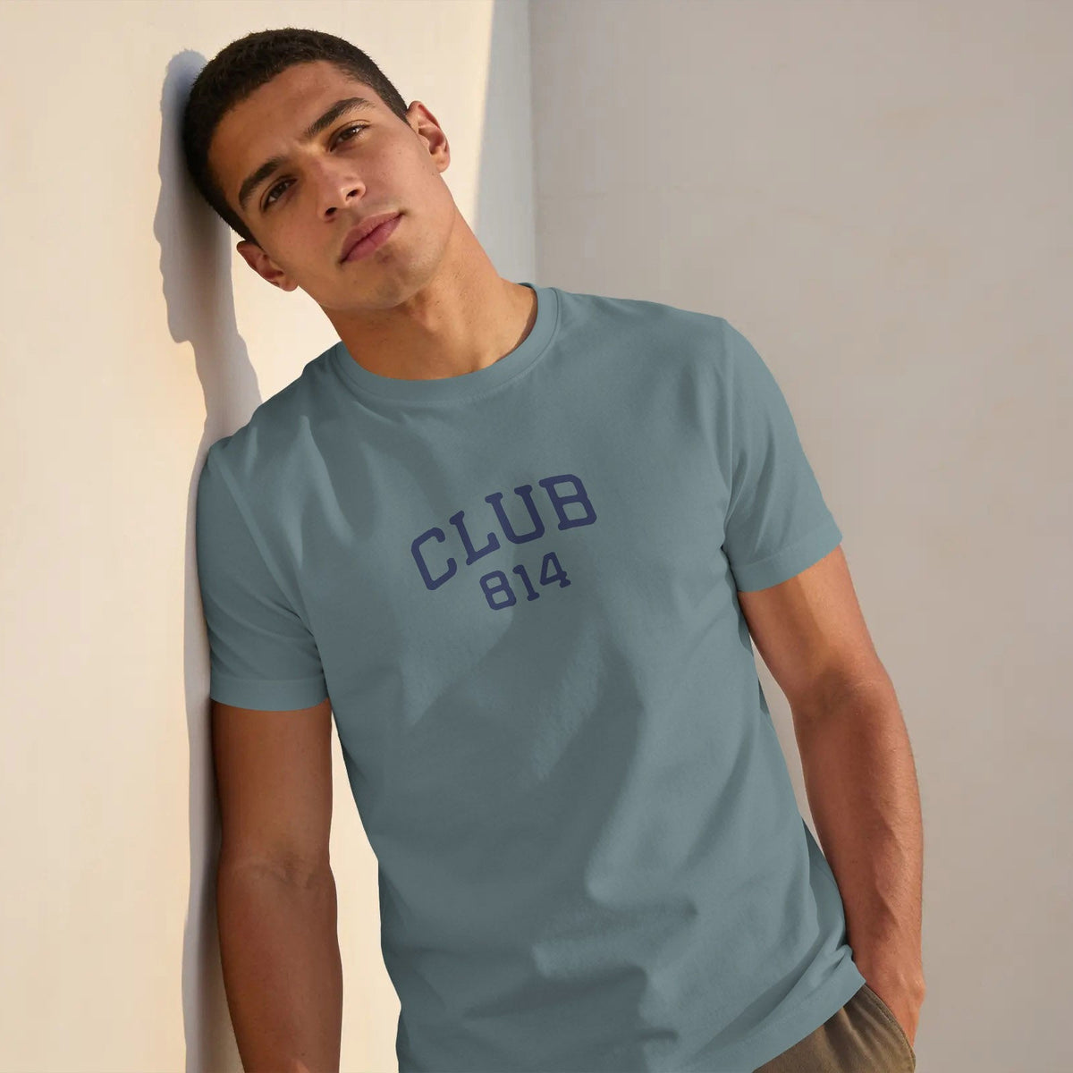 A man wearing a dusty blue crewneck t-shirt with 'CLUB 814' printed in navy blue on the chest, leaning against a beige wall.