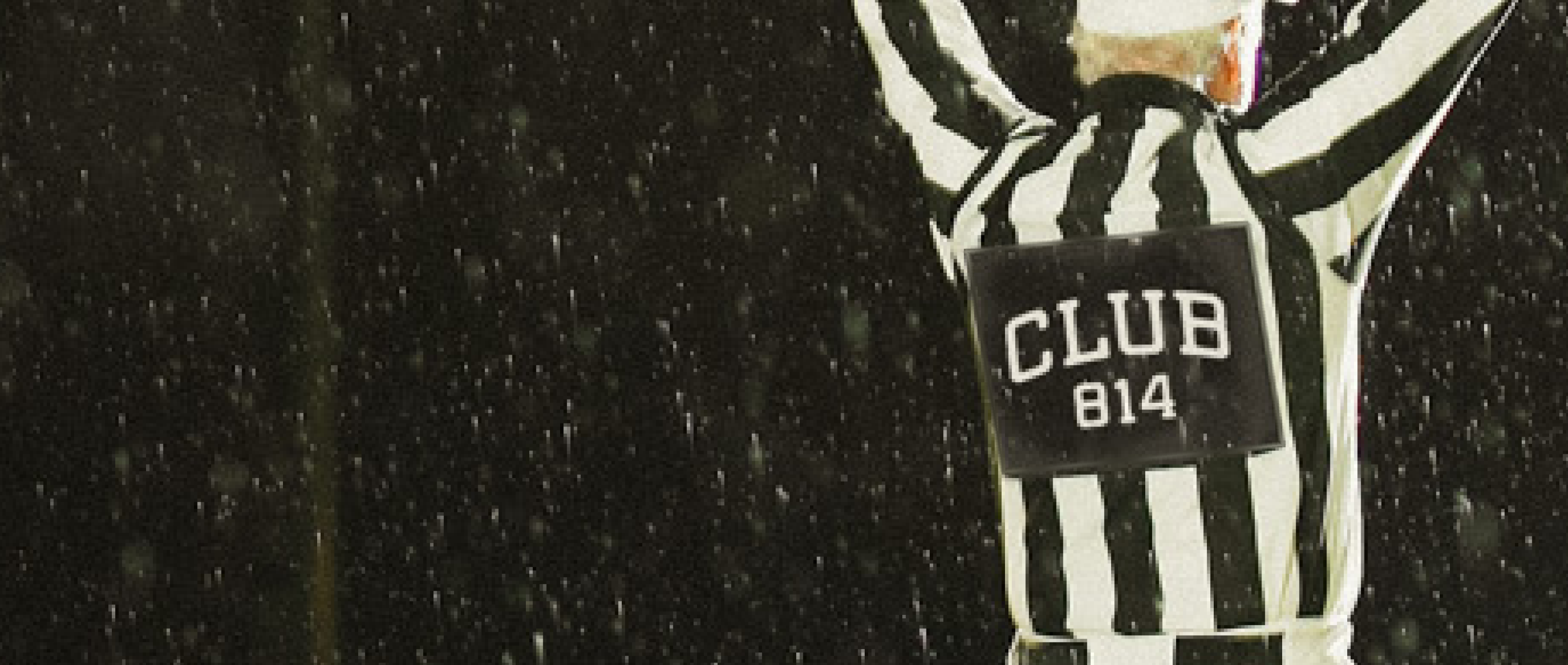 A referee in a black and white striped shirt with 'CLUB 814' on the back, raising both arms in the rain at night.