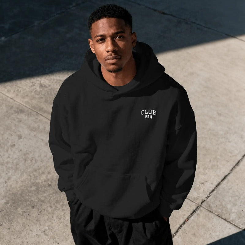 A man wearing a black pullover hoodie with a small white 'CLUB 814' logo on the left chest, standing outdoors on concrete pavement in shadowed sunlight.