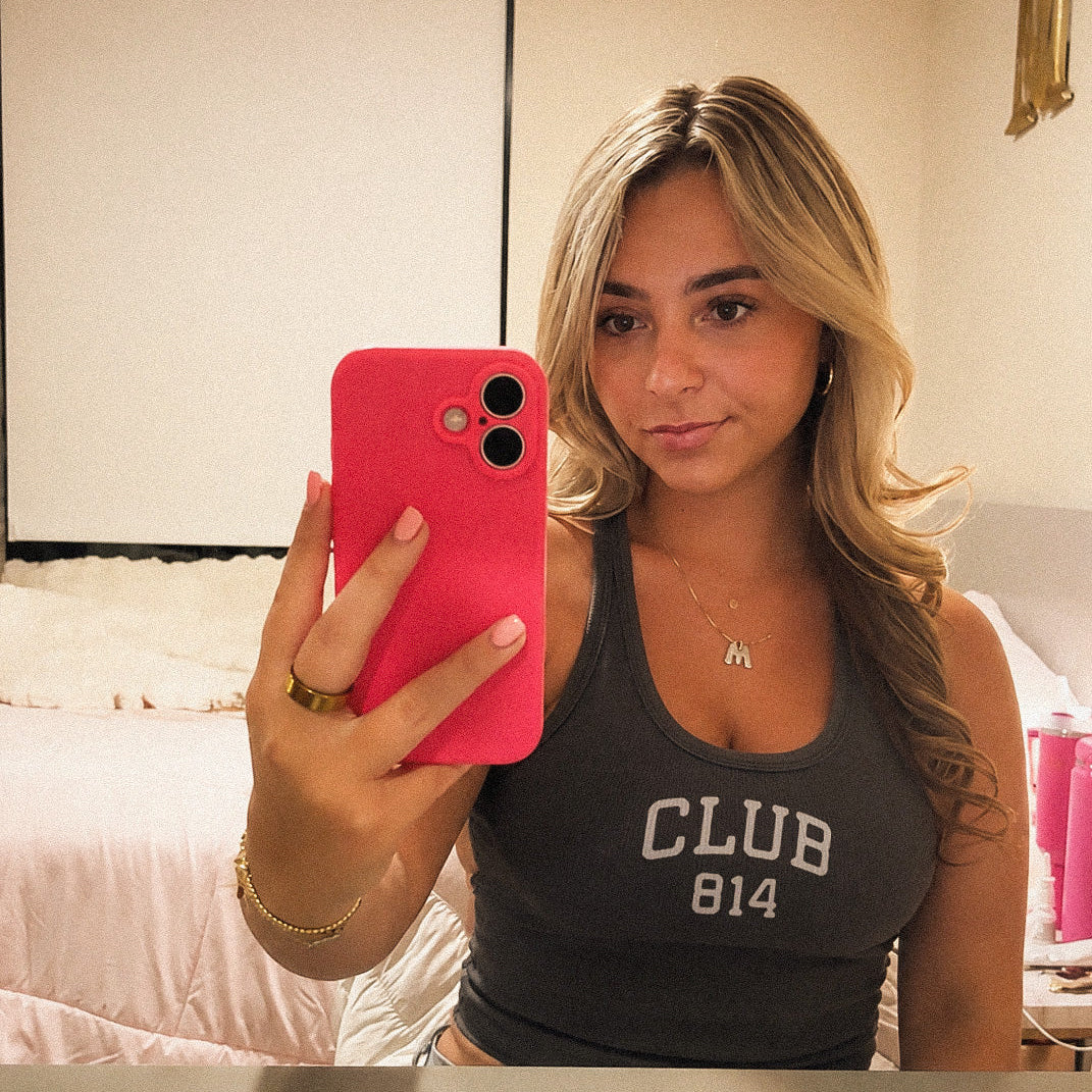 A woman taking a mirror selfie in a bedroom, wearing a dark gray tank top with 'CLUB 814' printed in white. She is holding a bright pink smartphone.