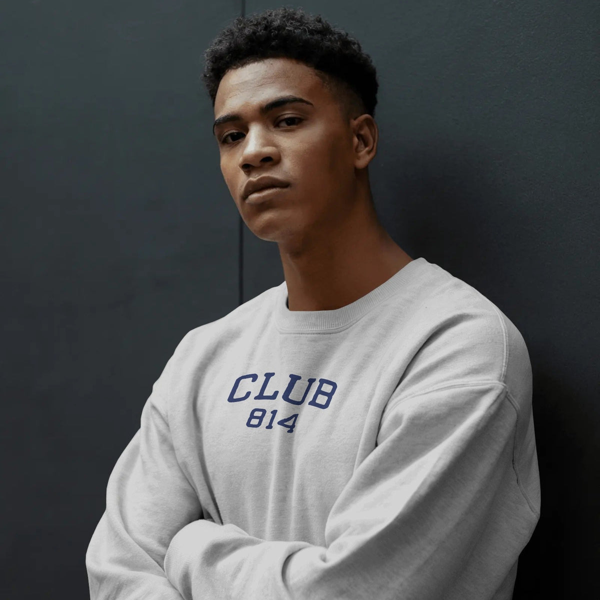 A young man with short dark curly hair wearing a light gray crewneck sweatshirt with 'CLUB 814' printed in navy blue on the chest, posing with arms crossed against a dark gray wall.