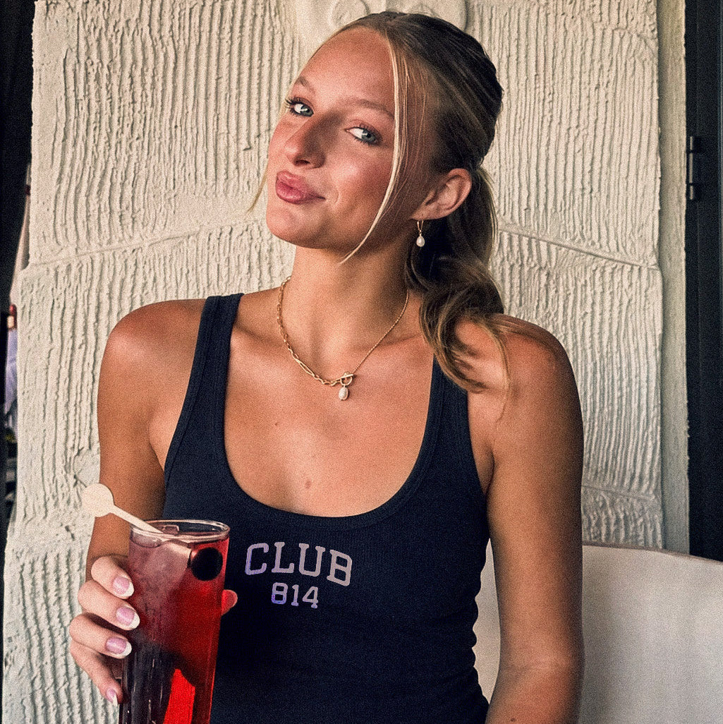 A woman wearing a black tank top with 'CLUB 814' printed in white, holding a glass of red cocktail in front of a textured beige wall.