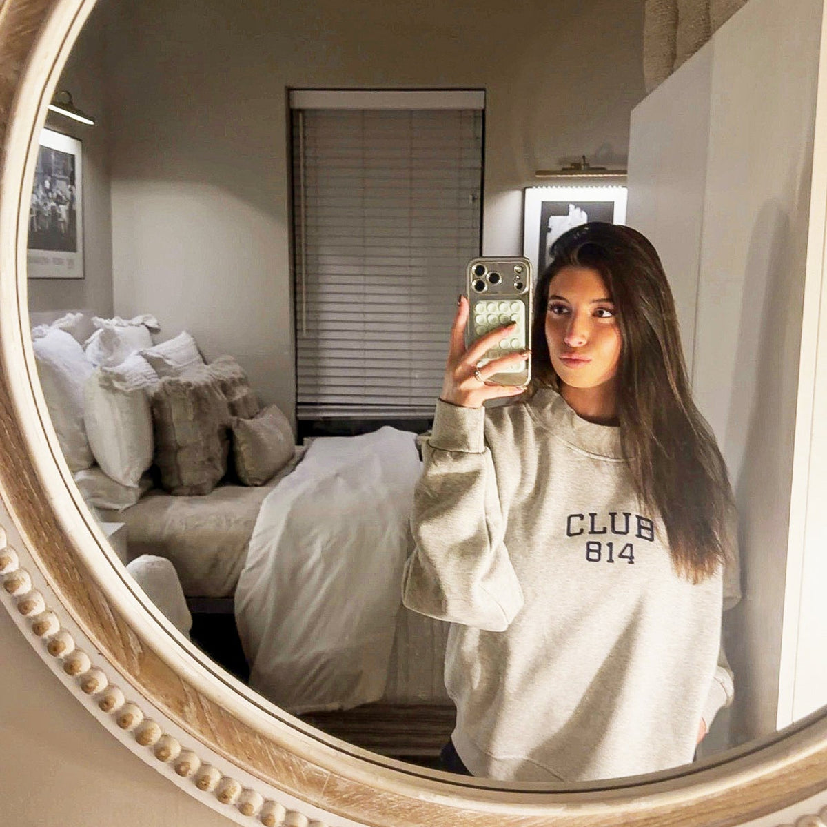 A woman taking a mirror selfie in a bedroom, wearing a light beige sweatshirt with 'CLUB 814' printed on the front. The image is framed by a large, ornate round mirror with a gold-toned beaded frame.