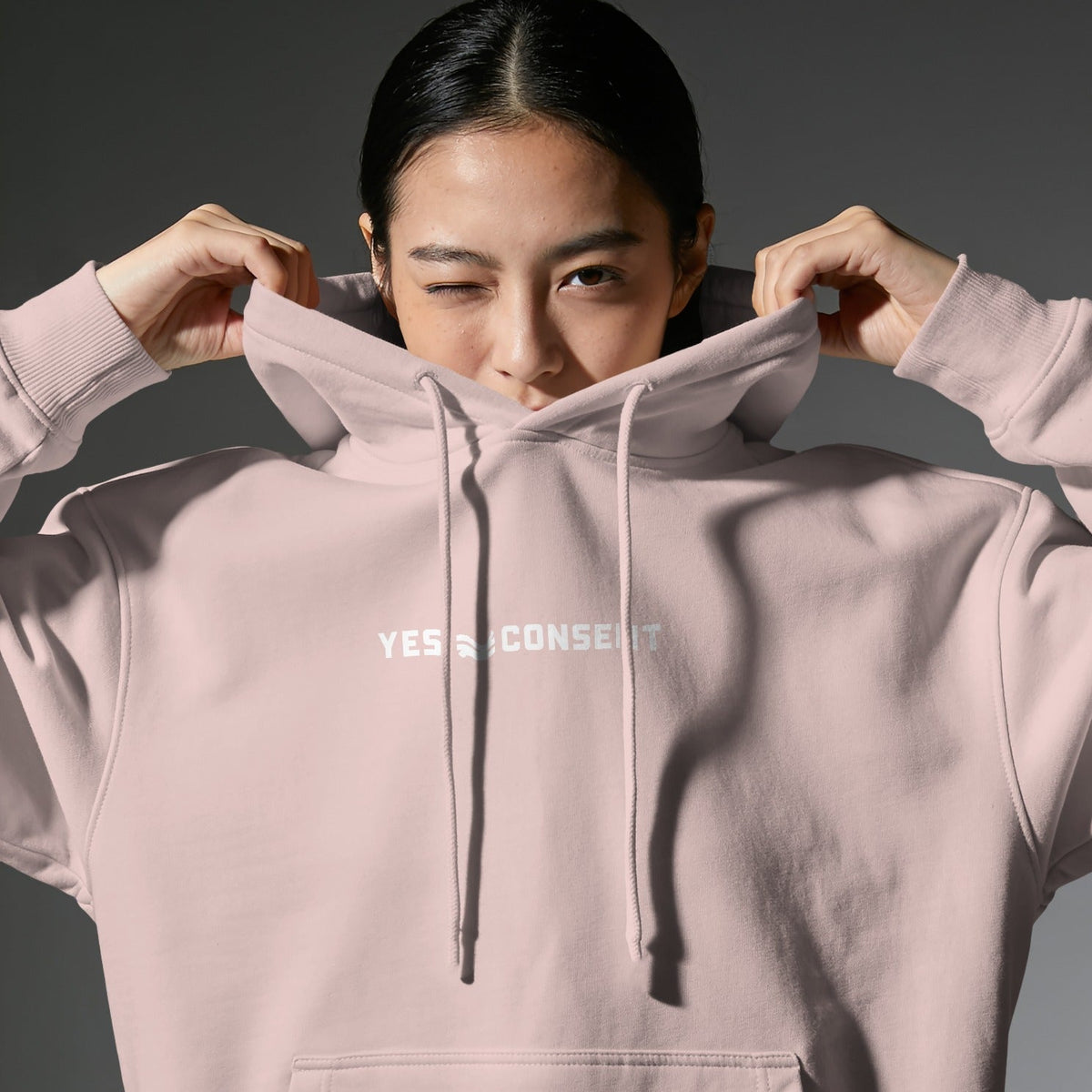 A model wearing a light pink pullover hoodie with the words 'YES ≠ CONSENT' printed in white across the chest, holding the hood up around her face against a dark gray studio background.